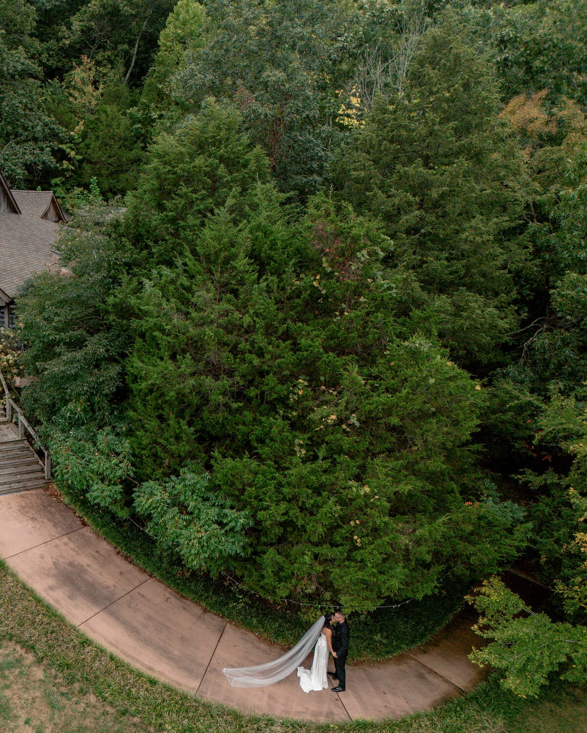 Bride and groom on a curving path beneath the trees at Chaumette Winery, photographed from above by drone, her veil trailing behind