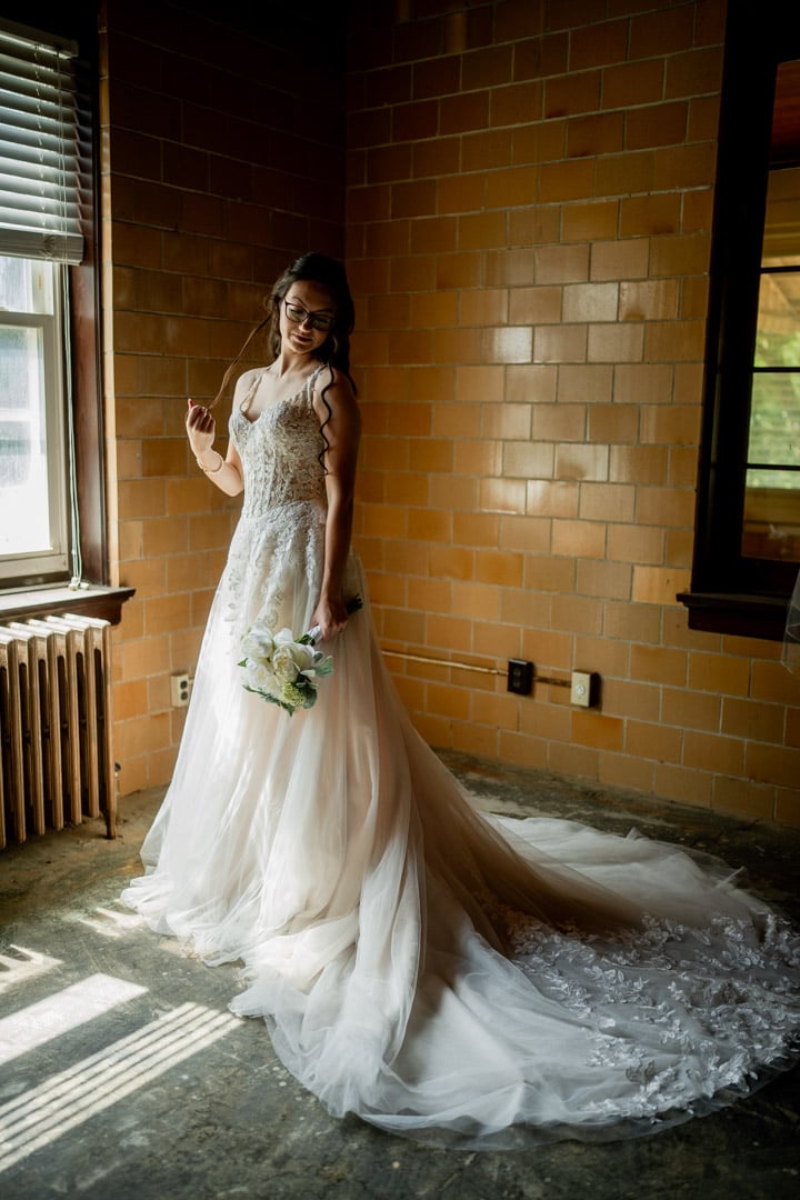 Bride in her wedding gown, softly lit by a window, holding her bouquet