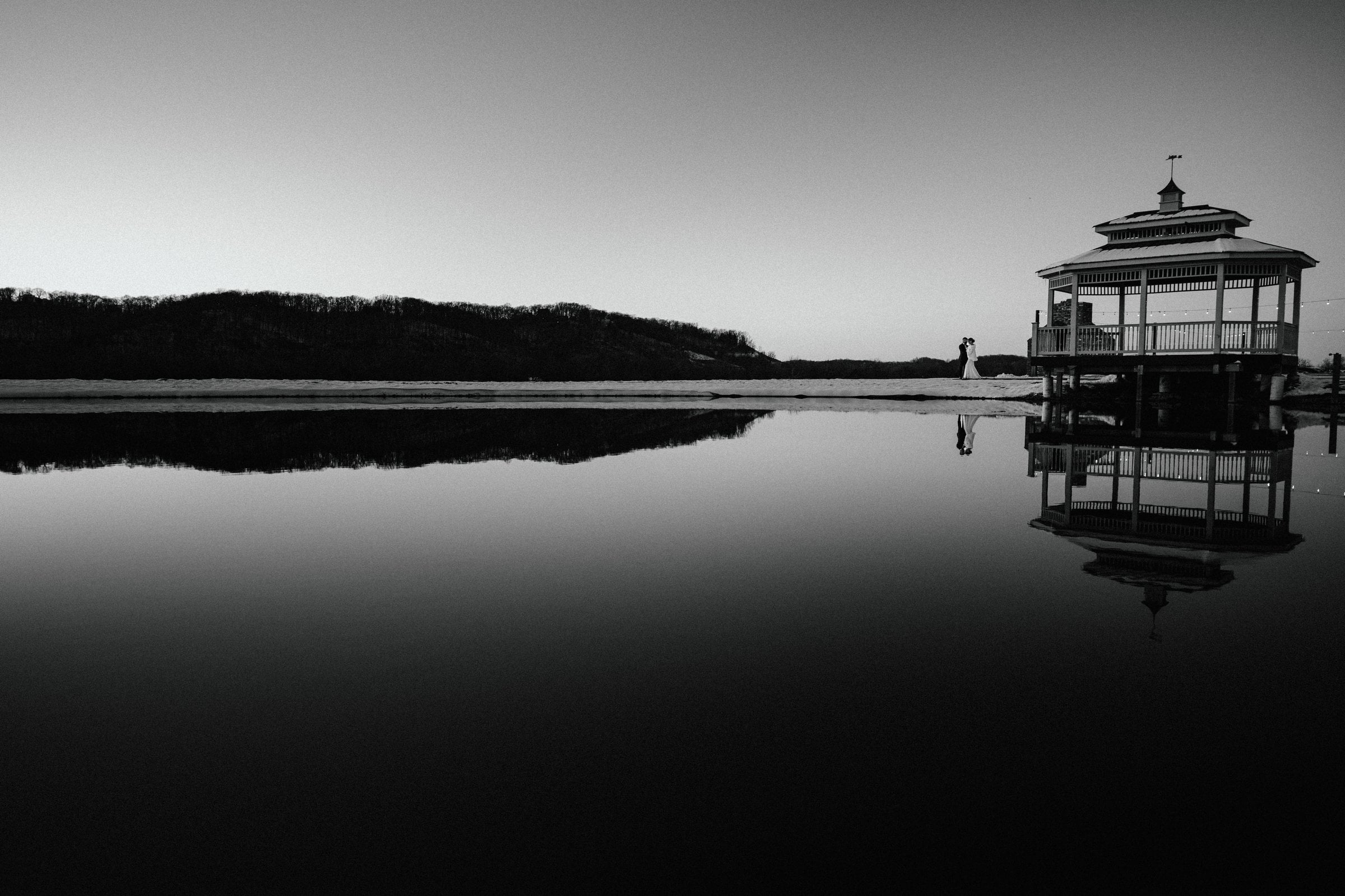 Black and white photo of a couple embracing at blue hour beside a lakeside gazebo, reflected in still water.