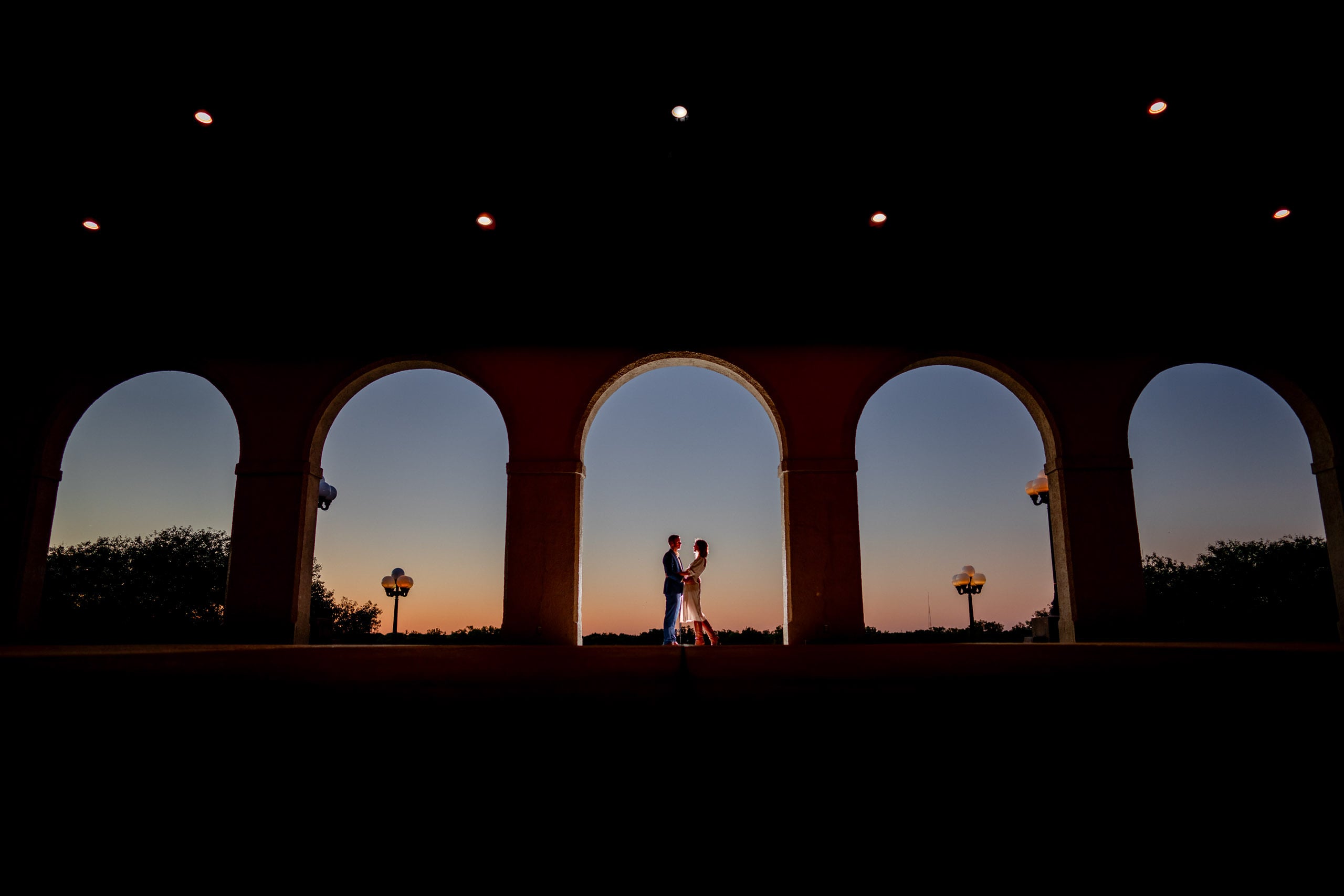 Silhouette of an engaged couple holding hands between tall arches at twilight