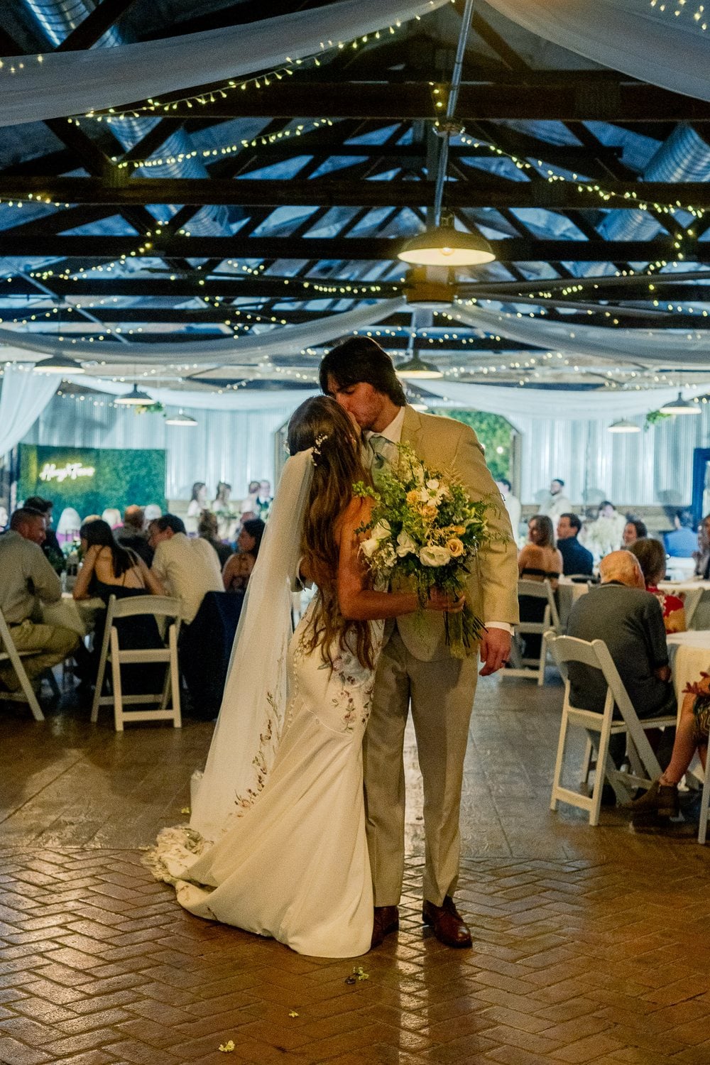Newlyweds kissing in the aisle of a string-light-lit barn venue as seated guests look on