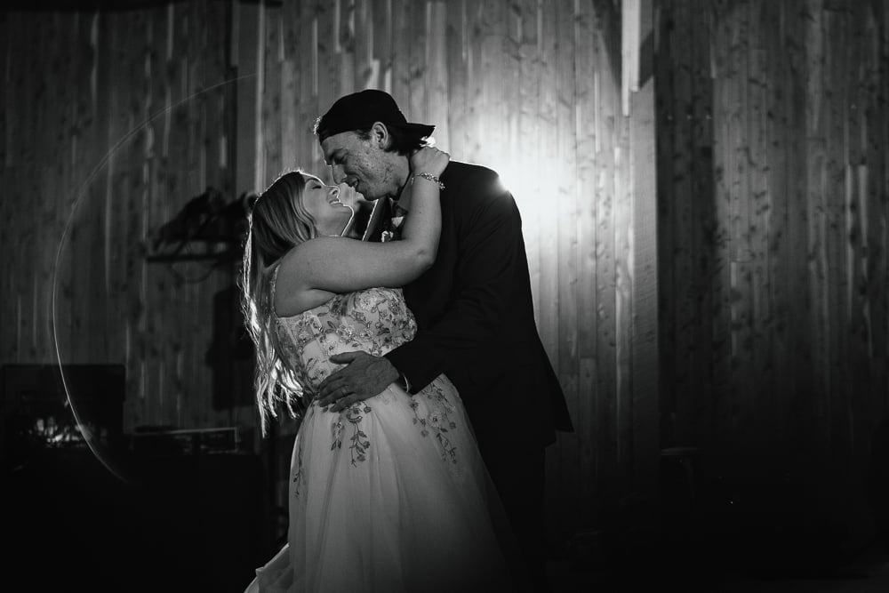 Black and white photo of a couple holding each other close during their first dance, backlit inside a barn