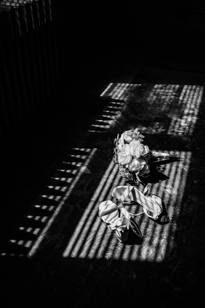 Black and white detail photo of a bridal bouquet and wedding shoes on a wood floor, lit by dramatic striped window-blind shadows