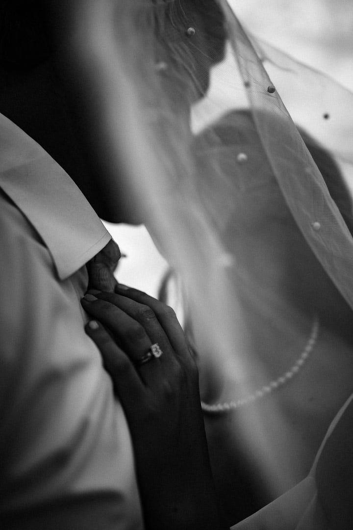 Black and white macro photo of a bride's hand and engagement ring resting on her partner's arm beneath her veil